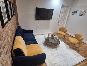 Ein Wohnzimmer mit einer Couch, zwei Sesseln, einem Glastisch, einem Fernseher und einer Tür.