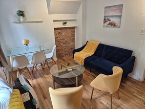 A modern living room with a glass table, white chairs, a blue sofa with a yellow throw, and a brick wall.