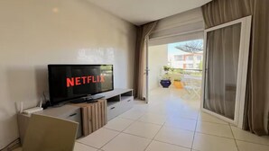 Un salon moderne doté d’un téléviseur à écran plat affichant le logo « NETFLIX », d’une porte-fenêtre coulissante donnant sur un espace extérieur et d’une table à manger avec des chaises.