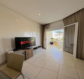 A modern living room with a flat-screen TV displaying 'NETFLIX,' a sliding glass door leading to an outdoor area, and a dining table with chairs. - Luxury Escape | Golf, Pools & Private Beach (Bouznika)