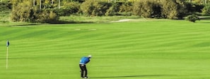 A golf course with a flagstick, a golfer in blue attire, and a backdrop of trees and bushes. - Luxury Escape | Golf, Pools & Private Beach (Bouznika)