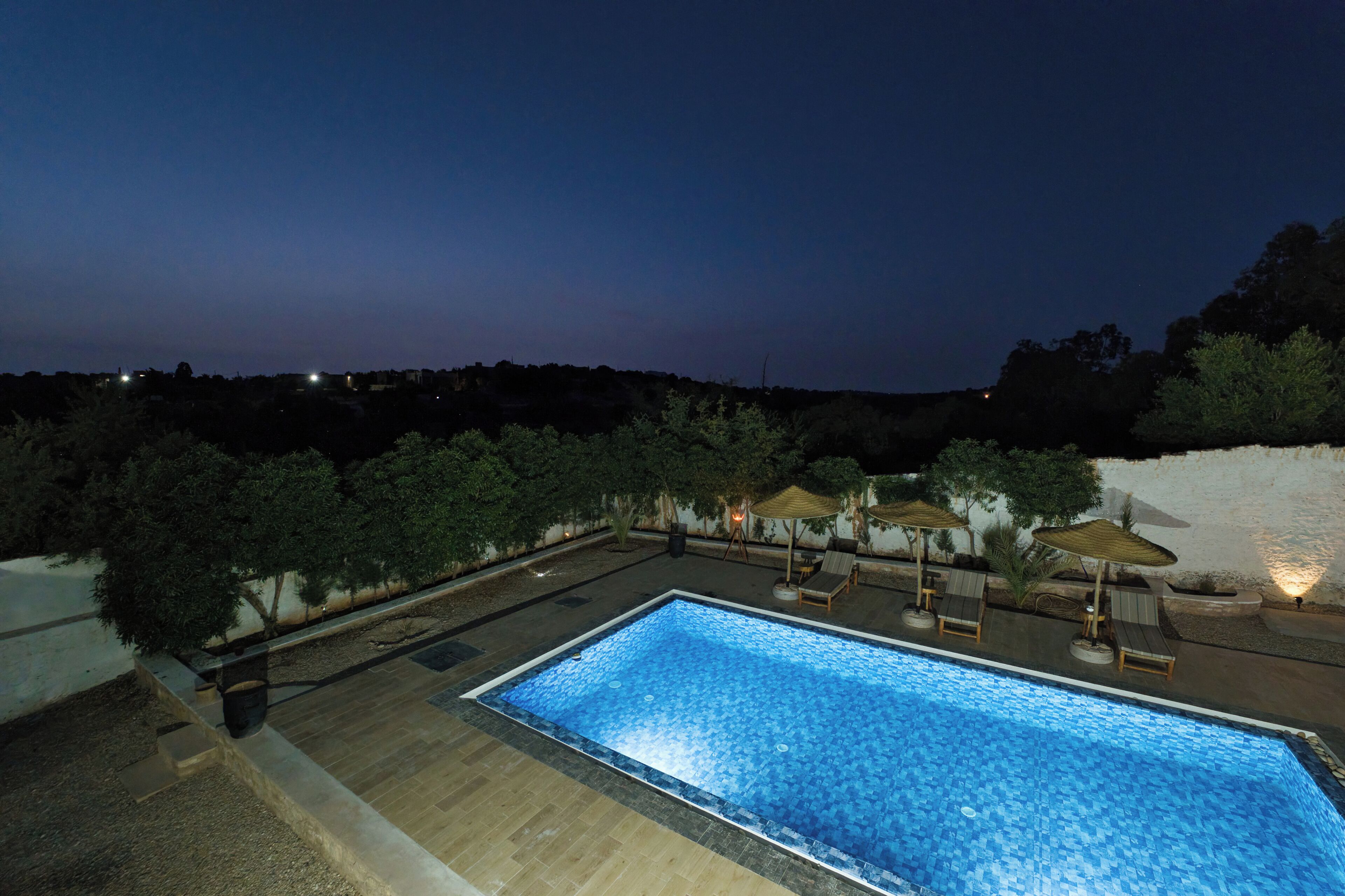 Une piscine rectangulaire bordée de carreaux, entourée de chaises longues et de parasols, se détachant sur un ciel nocturne.
