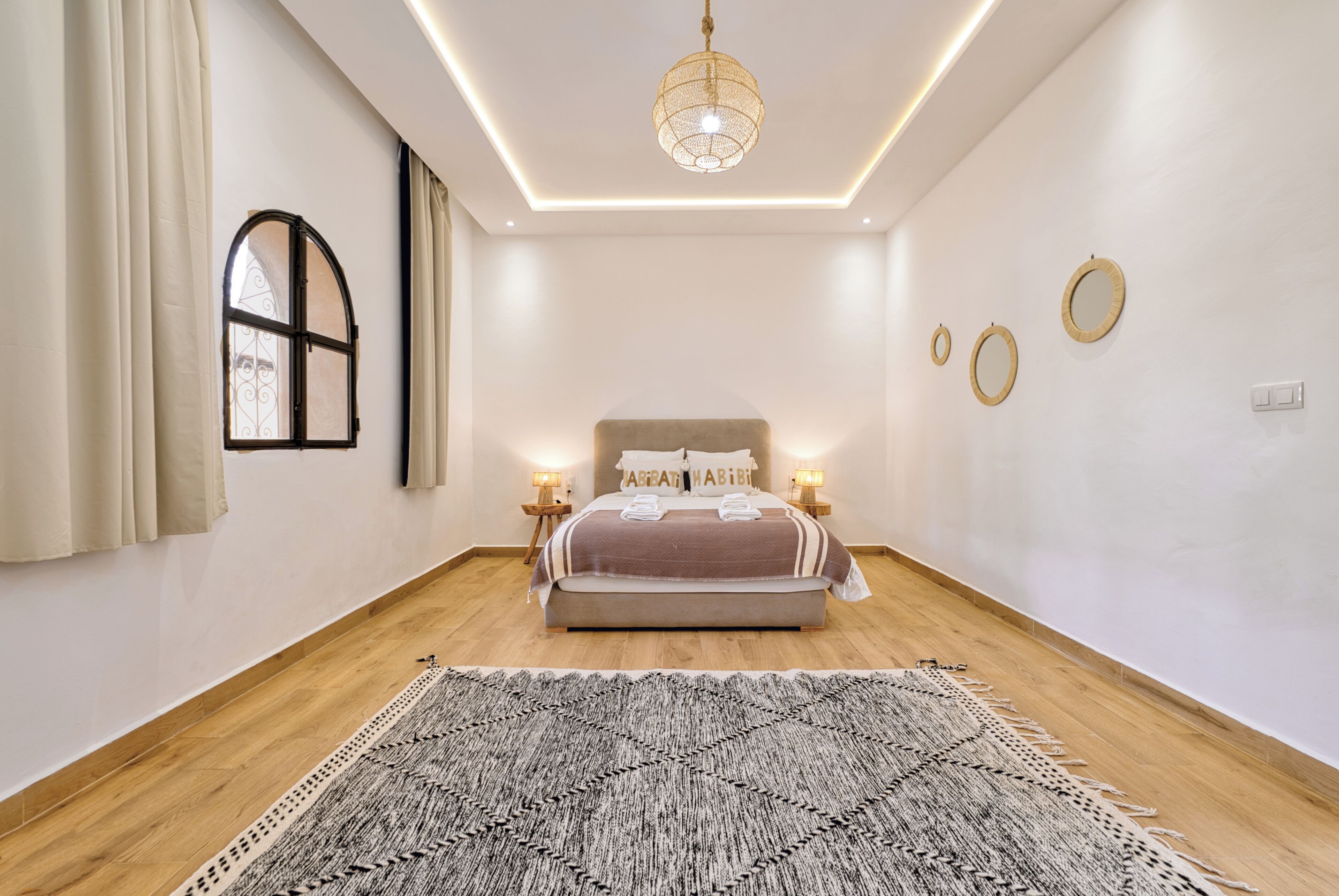 Une chambre à coucher avec un lit, des lampes de chevet, un luminaire suspendu, une fenêtre avec des rideaux et un tapis à motifs.