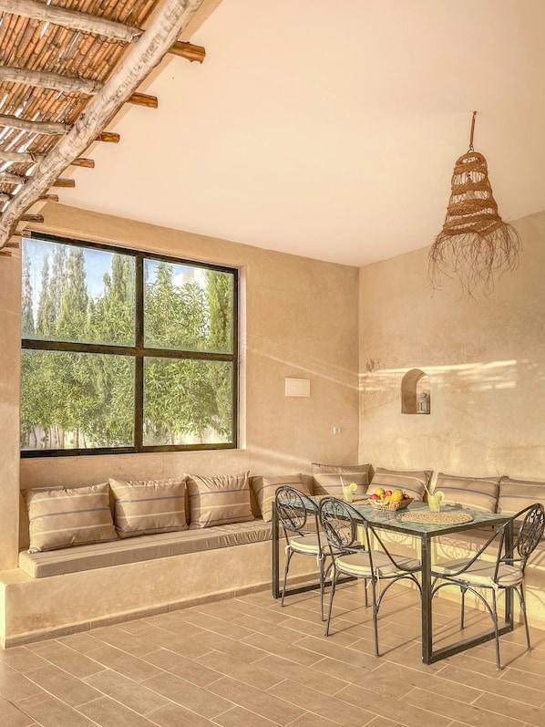 A room with a large window, a dining table with chairs, a hanging light fixture, and a built-in bench with cushions.