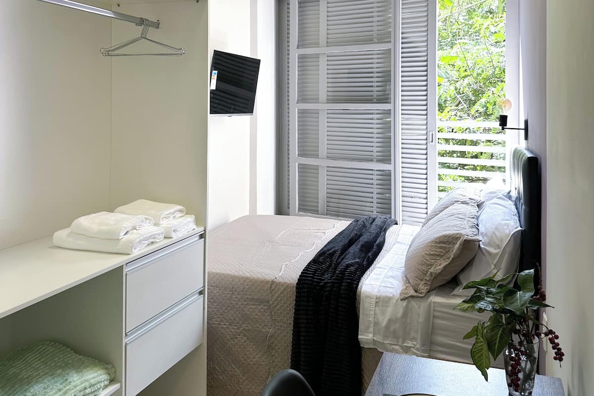 Une chambre avec un lit, un bureau avec une chaise, une fenĂȘtre avec des stores, une armoire et une petite plante sur le bureau.