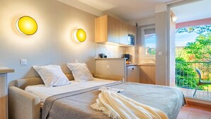 Een slaapkamer met een bed, nachtkastjes, een kleine kitchenette en een balkon met uitzicht op groen.