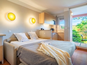 Une chambre avec un lit, des tables de chevet, une petite kitchenette et un balcon donnant sur un espace verdoyant.