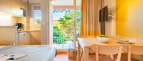 A hotel room with a bed, a dining table with chairs, a small kitchen area, and a balcony with a view of greenery.