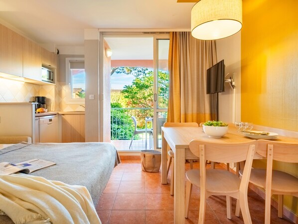 A hotel room with a bed, a dining table with chairs, a small kitchen area, and a balcony with a view of greenery.