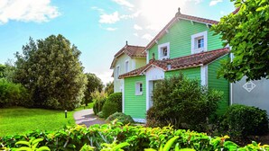 Ett grönt hus med ett tak täckt av tegelpannor, omgivet av frodig grönska och en klar himmel.