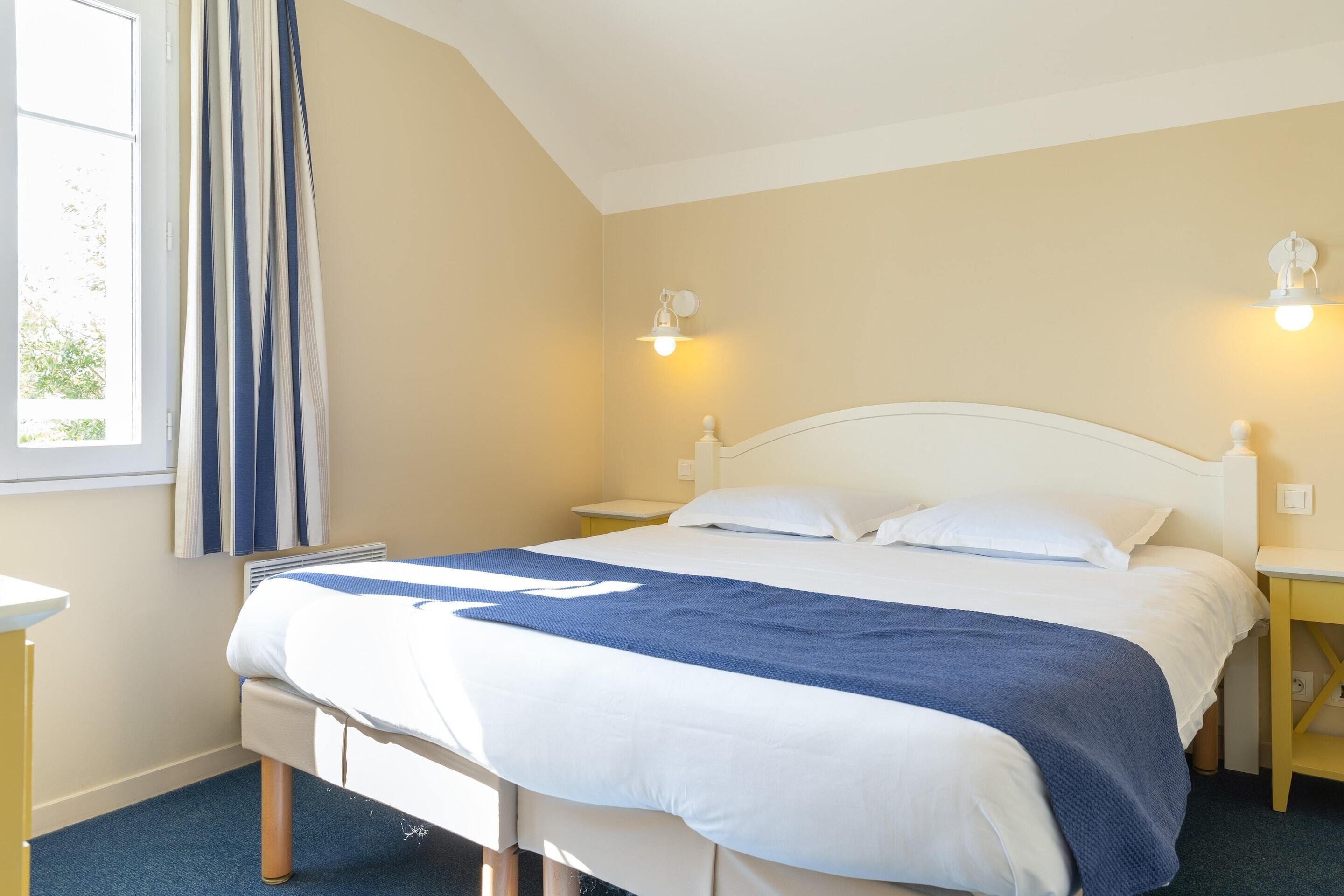 A neatly made bed with white and blue bedding, two bedside tables with lamps, a window with curtains, and a light-colored wall.