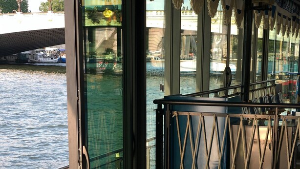 Un coin repas avec une longue table dressée pour deux personnes, un canapé à rayures et une vue sur l’eau grâce à de grandes fenêtres.