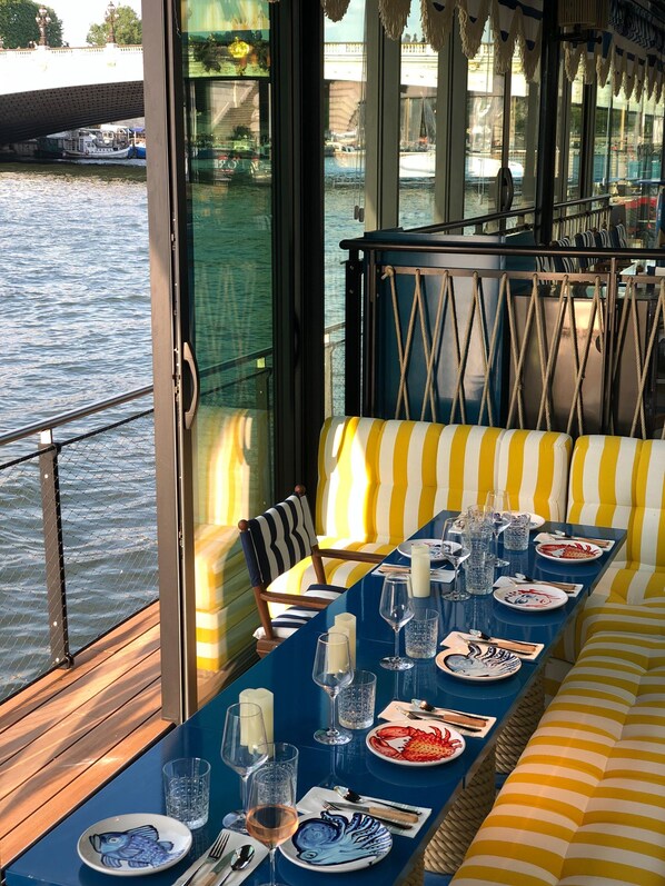 A dining area with a long table set for two, a striped sofa, and a view of the water through large windows.