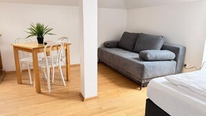 Avokeittiö-olohuone -tyylinen huone, jossa on puinen ruokapöytä, valkoiset tuolit, harmaa sohva ja sänky.