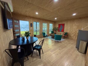 Ein geräumiges Zimmer mit Holzbalkendecke, großen Fenstern, einem Esstisch mit Stühlen, einer Couch und einem an der Wand befestigten Fernseher.