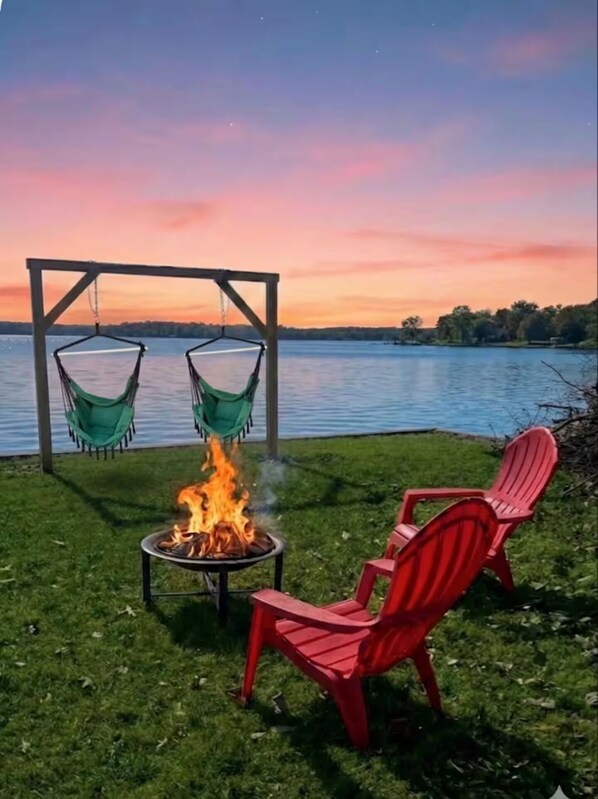 Ein Lagerfeuer mit zwei roten Stühlen und zwei grünen Hängematten unter einer Holzkonstruktion am See bei Sonnenuntergang.