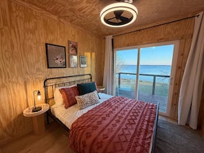 Une chambre dotée d’un grand lit, d’un ventilateur de plafond, d’une table de chevet avec une lampe, et offrant une vue sur la mer grâce à une porte-fenêtre coulissante.