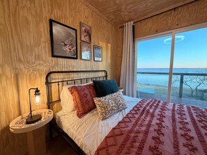 Une chambre à coucher avec un lit, une table de chevet, une fenêtre donnant sur la mer et des murs en bois.