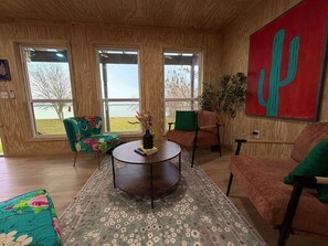 A room with a wooden ceiling, large windows, a round table with a vase, two chairs, a couch, and a cactus painting.
