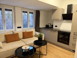 A modern living room with a sofa, a small round table, and a kitchen area with a stove and cabinets. - Faraway Homes Studio Semmering #308 (Spital am Semmering)