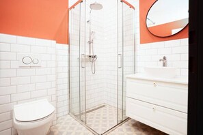 Ein Badezimmer mit einer gläsernen Duschkabine, weißen Fliesen, einer weißen Toilette und einem weißen Waschtisch mit Spiegel.