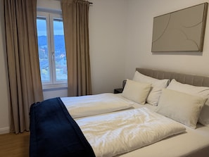 Un llit doble amb roba de llit blanca, una tauleta de nit, una làmpada i una finestra amb cortines.