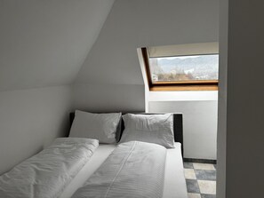 Ein kleines, modernes Schlafzimmer mit schrägen Wänden und einem Oberlicht, mit einem Bett mit weißer Bettwäsche und einem Holz-Kopfteil.