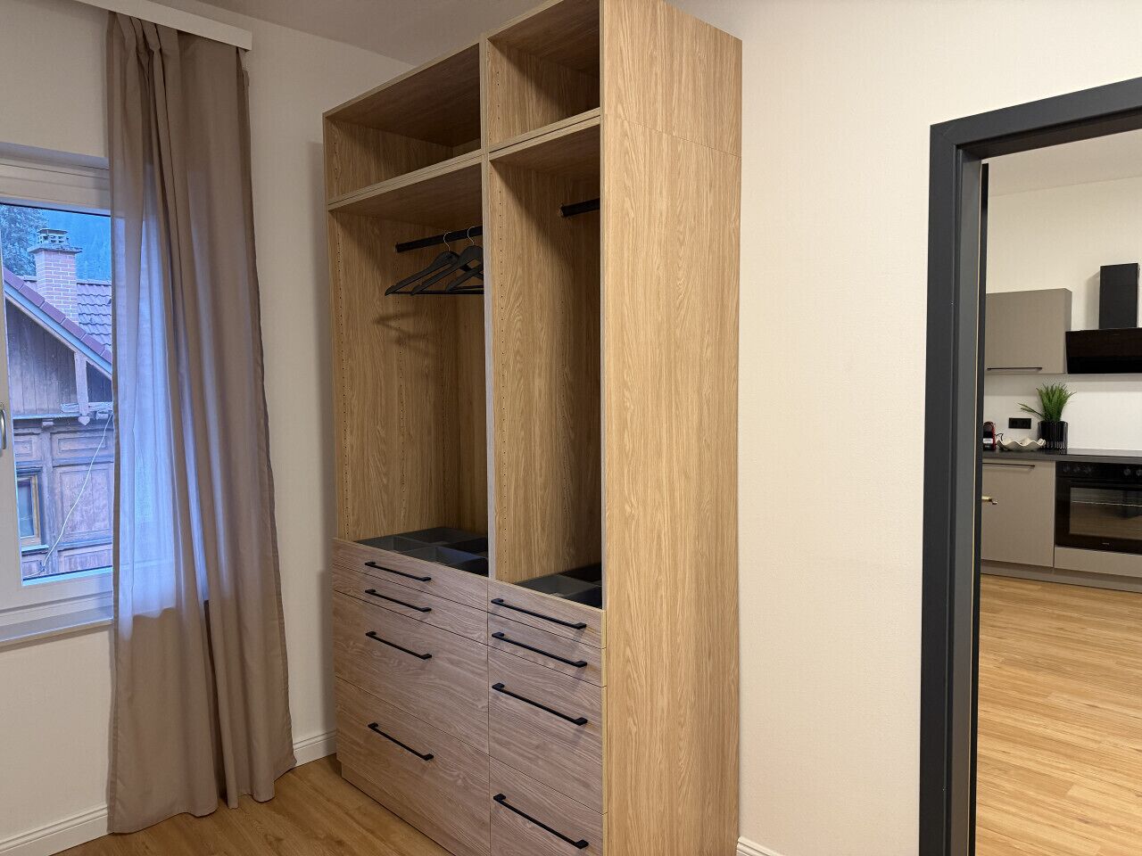 Een kamer met een houten garderobe, een raam met gordijnen en uitzicht op een huis.