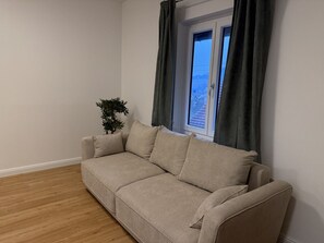 Un sofá color beige en una habitación con piso de madera y una ventana con cortinas.