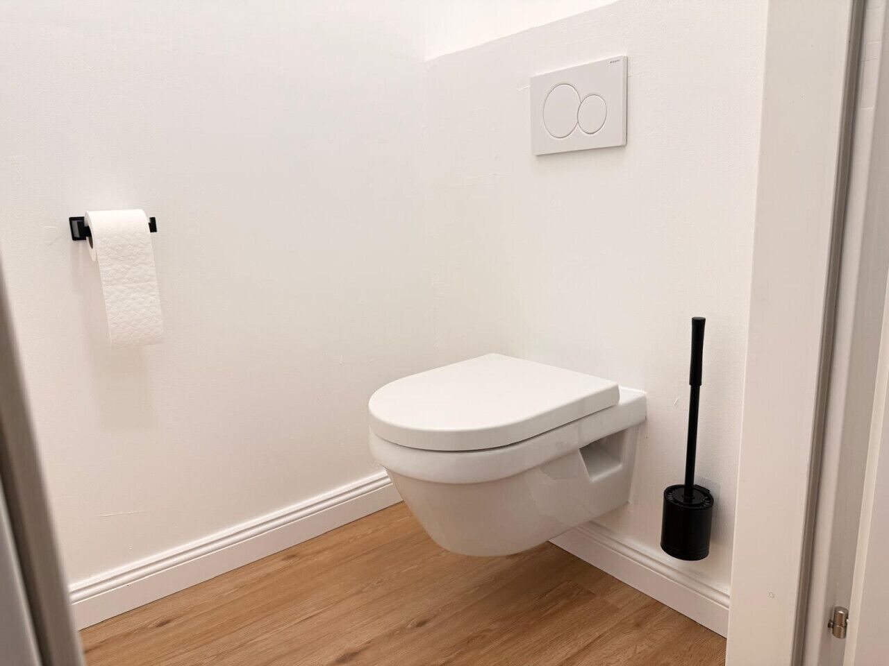 A white toilet with a flush handle, a toilet paper holder, and a black toilet brush on the floor.