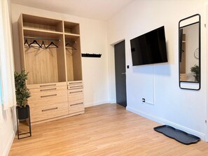 Ett rum med en garderob i trä, en svart klädhängare, en svart golvmatta, en svart TV som är monterad på väggen och en liten krukväxt.