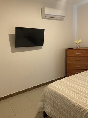 Ein Schlafzimmer mit einem Bett, einem Schminktisch, einem an der Wand montierten Fernseher und einer Klimaanlage.