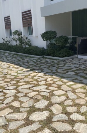 Ein gepflasterter Außenbereich mit Steinplatten und Gras, ein weißes Gebäude mit Fenstern und einer Tür sowie ein kleiner Baum.