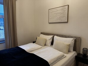 Ett sovrum med en säng, kuddar, ett sängbord, en lampa och ett fönster med gardiner.