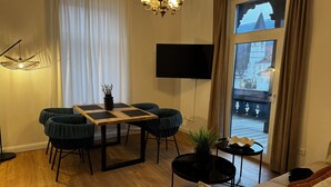 Een woonkamer met houten vloer, een eettafel met stoelen, een bank, een aan de muur bevestigde televisie en een groot raam met uitzicht op gebouwen.