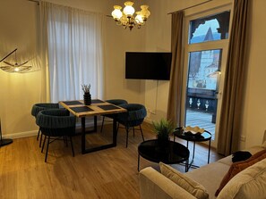 Ein möbliertes Zimmer mit Esstisch, Stühlen, einer Couch, einem Fernseher und Blick durch ein Fenster.