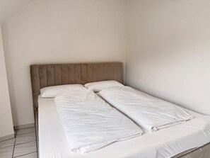 Ein Einzelbett mit Holz-Kopfteil in einem Raum mit schlichten Wänden und Fliesenboden.