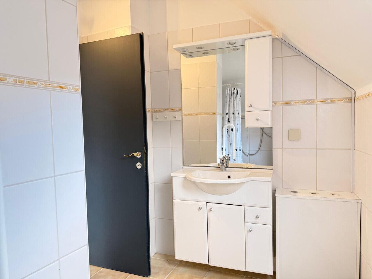 A bathroom with a white sink, mirror, and cabinets, a black door, and tiled walls.