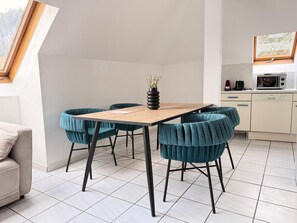 Ein moderner Essbereich mit einem Holztisch und stuhlbezogenen Sesseln in Türkis, einer Vase mit getrockneten Blumen und einer Küche im Hintergrund.