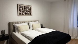 Een slaapkamer met een bed, een nachtkastje, een lamp en een kunstwerk aan de muur.
