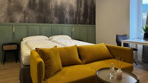 Een slaapkamer met een bed, een groen hoofdeinde, een mosterdkleurige bank, een klein tafeltje met een fles en een schilderij van bomen aan de muur.