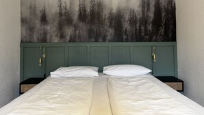 Een tweepersoonsbed met een groen hoofdeinde, twee nachtkastjes en een muurschildering met een bosmotief.