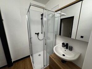 Ein modernes Badezimmer mit Duschkabine aus Glas, einem weißen Waschbecken mit zwei schwarzen Seifenspendern und einem Holz-Waschtisch.
