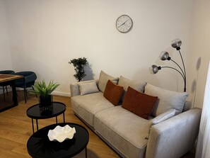 Ein Wohnzimmer mit einer Couch, einem runden Couchtisch, einer Zimmerpflanze, einer Wanduhr und einem Essbereich mit Stühlen und Tisch.