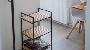 Um quarto com um radiador branco, uma mesa de jantar de madeira, uma cadeira branca, uma estante de metal preta com um livro e uma janela com cortinas florais.