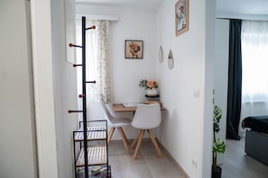 A small, well-lit room with a desk, two chairs, a coat rack, a framed picture, and a potted plant.