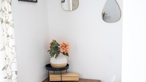 Uma área de refeições com mesa de madeira, cadeiras brancas, um vaso com flores e dois espelhos pendurados.