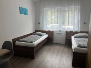 Ein kleines Zimmer mit zwei Einzelbetten, einem kleinen Schreibtisch und einem Fenster mit durchsichtigen Vorhängen.