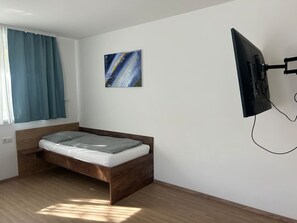Ein Einzelbett mit Holzrahmen, ein an der Wand befestigter Fernseher und ein Fenster mit Vorhängen.