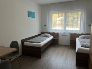 Habitación pequeña con dos camas, un escritorio y una ventana con cortinas translúcidas.
